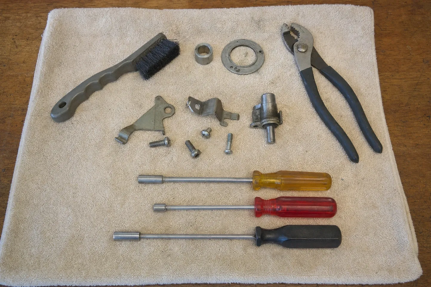Wire brush, slip-joint pliers, three nut drivers, a brass collar ring, a small valve body with a shaft and loose screws laid out in rows on a towel