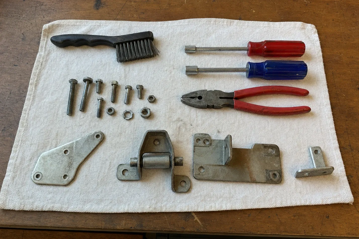 Four steel brackets including one hinge with a roller barrel, a row of bolts, nuts and washers, two nut drivers, pliers and a wire brush laid out on a cloth